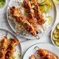 Plates of marinaded, sesame chicken kabobs, served on a bed of white rice with marinated cucumbers.